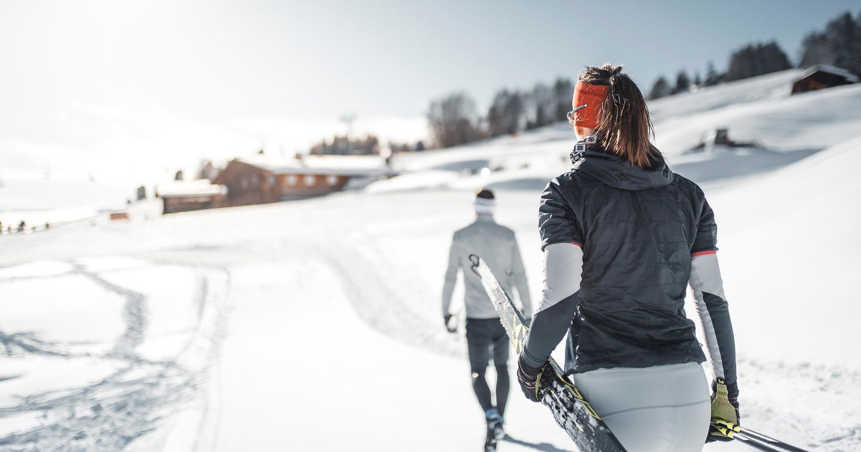 Zwei Langläufer gehen mit Skiern in der Hand durch eine verschneite Winterlandschaft, im Hintergrund Holzhütten und sanfte Hügel unter strahlend blauem Himmel