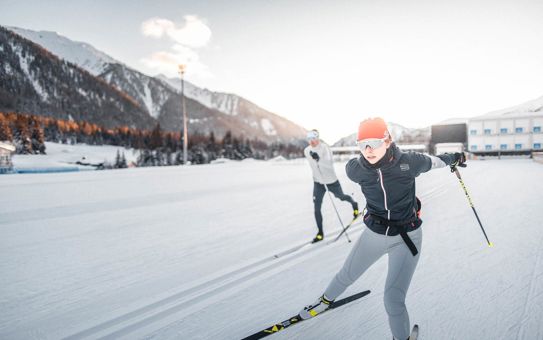 Two cross-country skiers training in the snowy biathlon arena in Antholz, a woman with a red headband in the foreground, snow-covered mountains in the background lit by the evening sun.