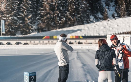 Biathlete shooting at the training range in Antholz, two other people talking nearby, snow-covered mountains in the background.