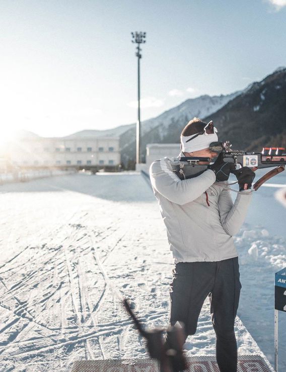 Biathlete aiming with rifle on the track in Antholz, watched by two people, snow-covered mountains and low sun in the background.