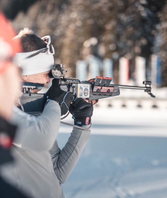 A biathlete aiming with a rifle at the shooting range on snow, with targets and flags in the background